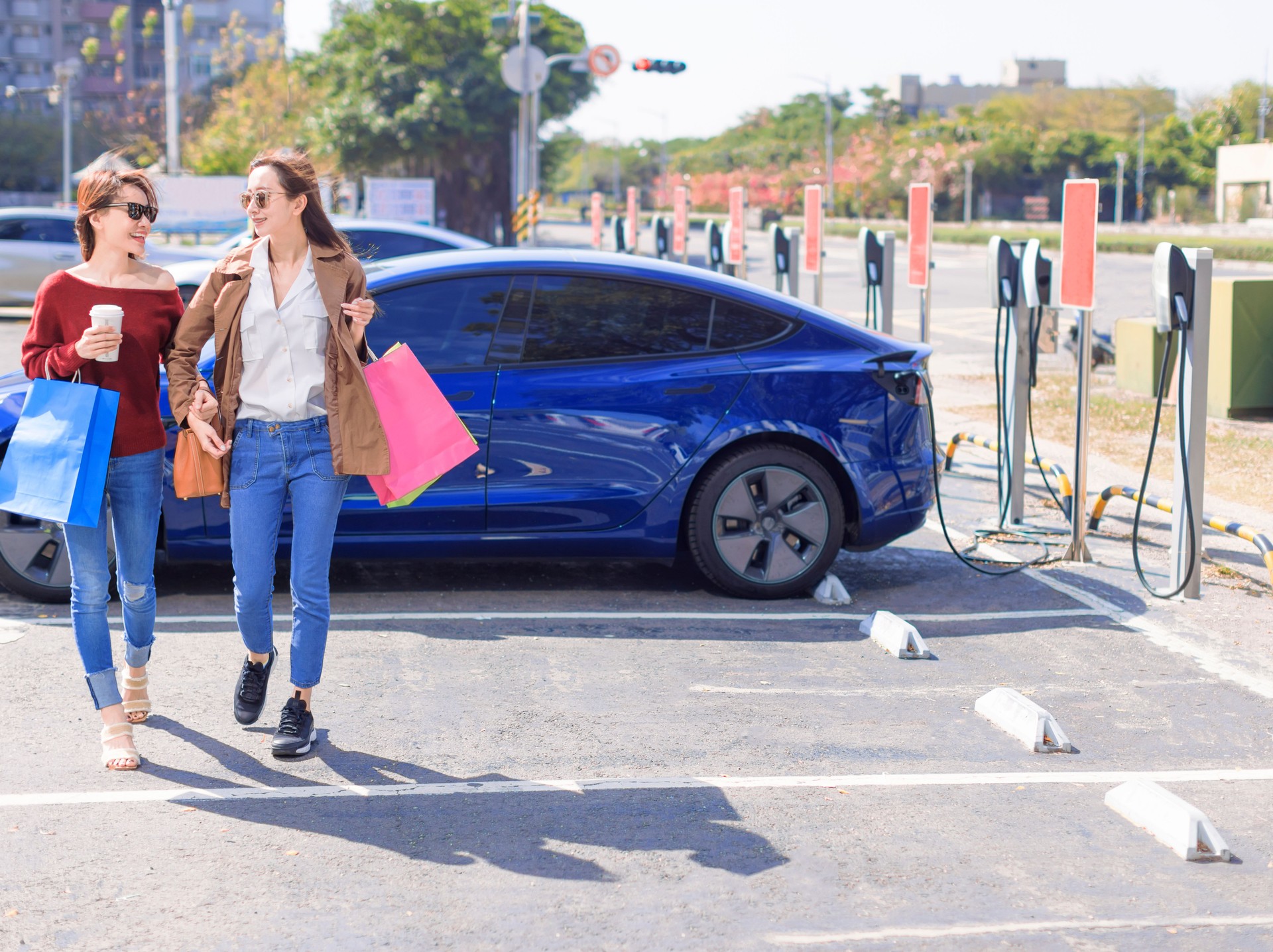 Happy  young woman standing on city parking near electric car, charging automobile battery from small city station, holding shopping bags and walking together